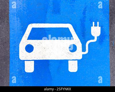 Eine Nahaufnahme zeigt ein weiß gemaltes Symbol eines Elektroautos, das an eine Ladestation angeschlossen ist und sich auf einem hellblauen Asphaltparkplatz befindet. Die Stockfoto