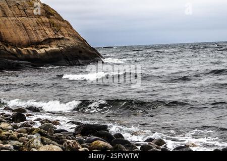 Wellen krachen sanft gegen glatte Kieselsteine entlang einer zerklüfteten Küstenklippe an der südnorwegischen Küste. Der bewölkte Himmel verstärkt die Ruhe. Stockfoto