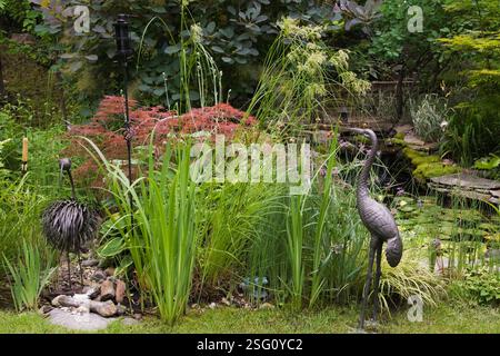 Strauß- und Reihvogelskulpturen aus Metall mit Acer palmatum inabe-shidare – japanischer roter Ahornbaum neben Teich mit Iris „gelbe Flagge“ im Garten Stockfoto