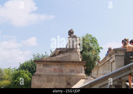 Eine Steinstatue einer sitzenden Figur, umgeben von Grün und Menschen auf einer Treppe. Die Statue ist detailliert und scheint Teil eines größeren Archits zu sein Stockfoto