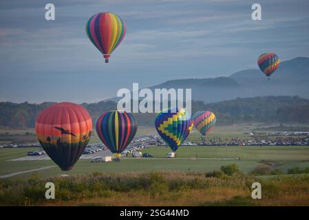Beim Adirondack Balloon Festival schweben bunte Heißluftballons über einer üppigen Landschaft mit bergiger Kulisse. Stockfoto