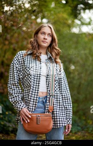 Herbstmodeporträt: Stilvolle junge Frau in schwarz-weißem kariertem Hemd und blauer Jeans posieren mit Tan-Leder-Umhängetasche im Naturpark. Lässig Stockfoto
