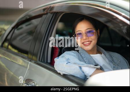 Eine attraktive asiatische Frau in Jeansjacke und Sonnenbrille sitzt in ihrem Auto und lächelt in die Kamera. Transport, Lebensstil, Reisen, Freiheit Stockfoto