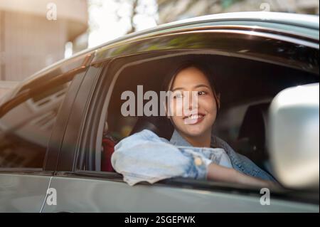 Eine attraktive, positive asiatische Frau in einer Jeansjacke sitzt in ihrem Auto, blickt aus dem Fenster und blickt während der Fahrt auf die Aussicht. Transport, Lebensstil Stockfoto