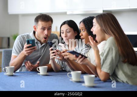 Erstaunte interrassische Gruppe von Freunden, die zu Hause Telefone in der Küche checken Stockfoto
