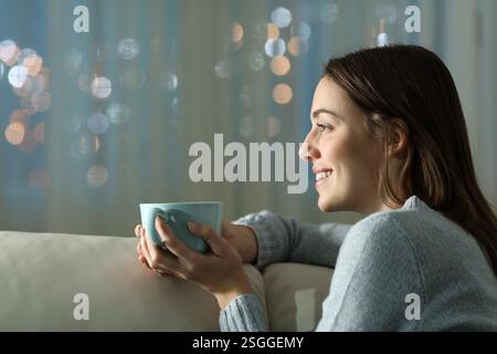 Glückliche Frau in der Nacht zu Hause, die Kaffee trinkt, auf einer Couch sitzt und wegblickt Stockfoto