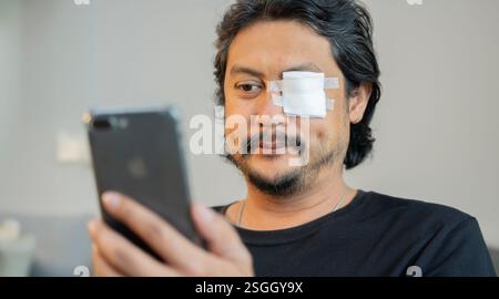 Erwachsener asiatischer Mann mit Augenschmerzen, bedeckt von einem Verband, der auf dem Sofa sitzt und sein Smartphone benutzt. Selektiver Fokus. Stockfoto