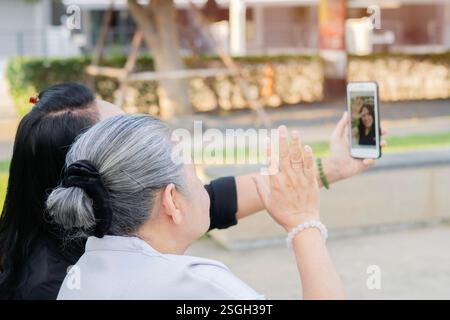 Glückliche asiatische Rentnerin, die mit ihrer Tochter im Park auf der Bank sitzt und mit der Familie Videoanrufe über das Handy nutzt. Stockfoto