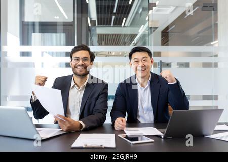 Porträt zweier Geschäftsleute, Männer, die glücklich in die Kamera blicken, den Sieg und den erfolgreichen Abschluss des Projekts feiern, während sie am Schreibtisch sitzen, in die Kamera schauen. Stockfoto