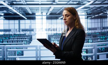 Computerwissenschaftler, der die Überprüfung durchführt und Daten auf Tablets mit den erforderlichen Fehlerbehebungen im Server-Hub eingibt. Frau, die im Rechenzentrum arbeitet und eine fortschrittliche Datenspeicherinfrastruktur zur Unterstützung von IT-Workloads bietet, Kamera B Stockfoto