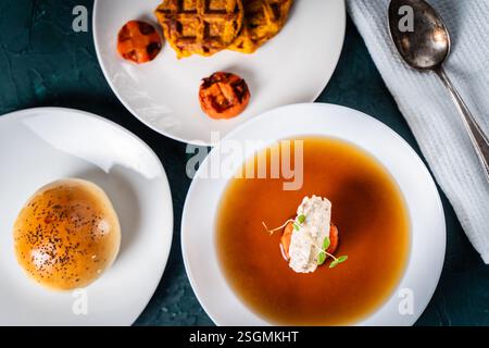 Suppe oder Consomme, dicke Fleischbrühe mit Karotten, Hähnchenknödel serviert auf einem weißen Teller auf dunkelgrünem Hintergrund mit Sesambrötchen, Serviette und Stockfoto