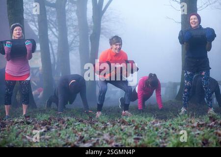 Eine Gruppe von Erwachsenen, die im Freien in einem nebeligen Wald trainieren, gewichtete Sandsäcke tragen und bunte Sportkleidung tragen. Sale Water Park, Manchester, Großbritannien Stockfoto