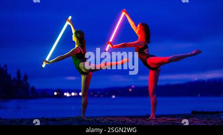 Zwei Tänzerinnen mit Neonlichtschrauben spielen ausdrucksstarke Posen am Strand in der Abenddämmerung, USA Stockfoto