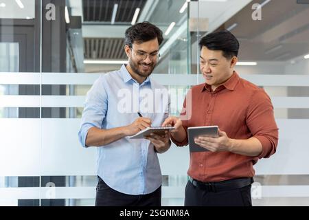 Zwei lächelnde und freudige Kollegen stehen im Büro, die über das aktuelle Arbeitsprojekt sprechen. Geschäftsleute in lässiger Kleidung reden. Stockfoto