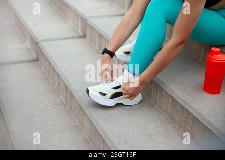 Im Freien sitzt eine Person in lebendiger Trainingskleidung auf Betonstufen und passt ihre Schnürsenkel an, bevor sie eine Trainingseinheit beginnt Stockfoto