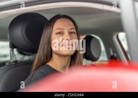 Eine junge Frau mit langen braunen Haaren und einem selbstbewussten Lächeln sitzt auf dem Fahrersitz eines modernen Autos Stockfoto