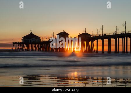 Das HSlow-Motion-Bild des Untington Beach Piers in Kalifornien zeigt die untergehende Sonne, die Licht durch die Pfeiler wirft, bevor es hinter dem Horizont untergeht. Stockfoto