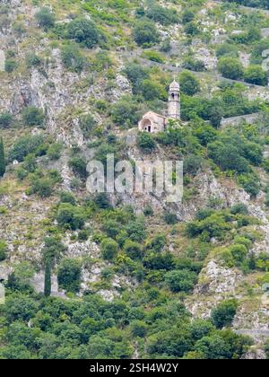 Kotor, Montenegro - 2. Juli 2024: Kirche unserer Lieben Frau von Remedy auf dem hohen grauen Berghang über der historischen Altstadt. Flecken von grünen Büschen Stockfoto