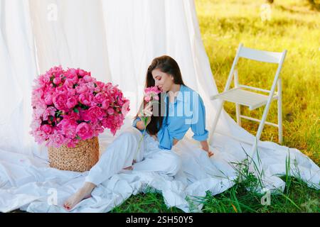 Eine stilvolle Frau in einem blauen Hemd und einer weißen Hose sitzt draußen barfuß auf einem weißen Stoff und riecht nach rosa Pfingstrose. Ein großer Korb mit Blumen und ein Weißer Stockfoto