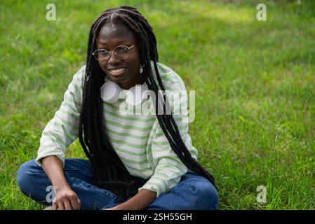 Porträt einer glücklichen jungen schwarzen Frau mit Dreadlocks, die Kopfhörer um den Hals und eine Brille tragen, die im Kreuz auf grünem Gras sitzt und einen Moment der Ruhe und Freizeit genießt Stockfoto