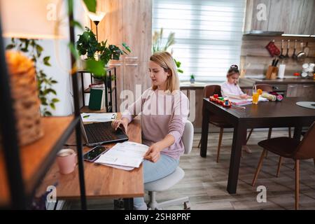 Frau, die am Laptop arbeitet, während Kind kreativ in der gemütlichen Küche spielt Stockfoto