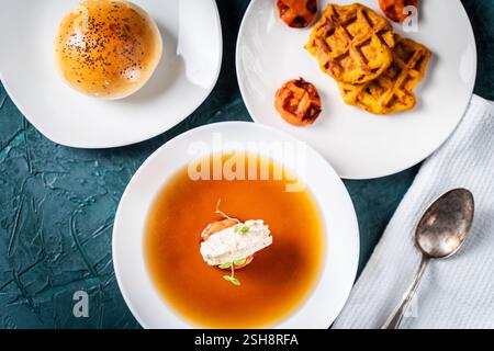 Suppe oder Consomme, dicke Fleischbrühe mit Karotten, Hähnchenknödel serviert auf einem weißen Teller auf dunkelgrünem Hintergrund mit Sesambrötchen, Serviette und Stockfoto