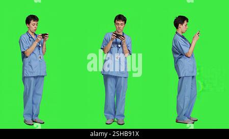Medizinische Assistentin spielt mobile Videospiele auf Smartphone-App und macht eine Pause von der medizinischen Arbeit. Schwester liebt das Gaming-Turnier am Telefon und steht vor Greenscreen-Hintergrund. Kamera A.. Stockfoto