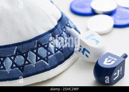 Das enge Bild eines weißen und blauen Yarmulke mit Dreidels und silbernen und blauen Münzen für die Hanukkah-Feier auf weißem Holzhintergrund Stockfoto
