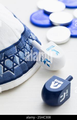 Das enge Bild eines weißen und blauen Yarmulke mit Dreidels und silbernen und blauen Münzen für die Hanukkah-Feier auf weißem Holzhintergrund Stockfoto