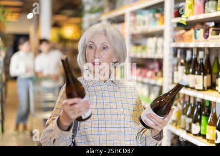 Senior weibliche Kundin im Supermarkt prüft Flaschen Wein, wählt alkoholische Getränke. Stockfoto