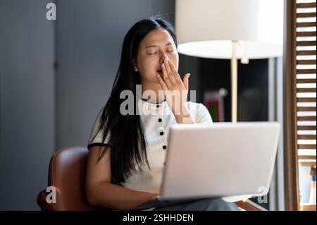 Eine müde asiatische Frau bedeckt ihren Mund während sie gähnt, während sie in einem Café an ihrem Laptop arbeitet und sich langweilig oder überarbeitet fühlt. Stockfoto