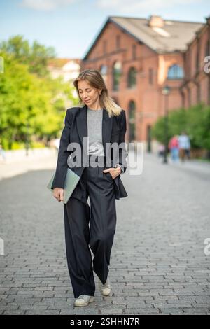 Lächelnde, coole Geschäftsfrau, die mit einem Laptop im Freien spaziert und unterwegs Arbeit und Leben in Einklang bringt Stockfoto
