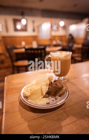 Blick auf das Innere des Cafés auf Apple Pie a la Mode und Apfelwein in einer Glasschale mit Schlagsahne und Karamellsauce auf einem Holztisch. Stockfoto