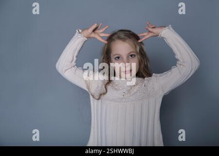 Ein Mädchen mit langen Haaren hebt spielerisch die Hände in einer selbstbewussten Pose und trägt einen stylischen Pullover. Dieses Foto zeigt sowohl ihre lustige Persönlichkeit als auch die Stockfoto
