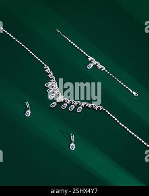 Kunstvoll verzierte Silberschmuck-Set: Halskette, Ohrringe, Armband mit blauen Tropfen-Edelsteinen und Diamanten auf grünem Hintergrund mit dramatischer diagonaler Beleuchtung Stockfoto