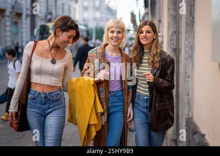 Eine Gruppe von drei modischen jungen Frauen spaziert lächelnd und lachend durch eine belebte Straße der Stadt. Ihr fröhlicher Ausdruck, trendige Outfits und Casua Stockfoto