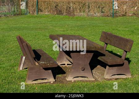 Ein rustikaler Picknicktisch aus Holz steht auf einem üppig grünen Rasen in einem Park. Die Sonne scheint hell und wirft sanfte Schatten auf den Boden. In der Nähe befindliche Büsche bieten einen Stockfoto
