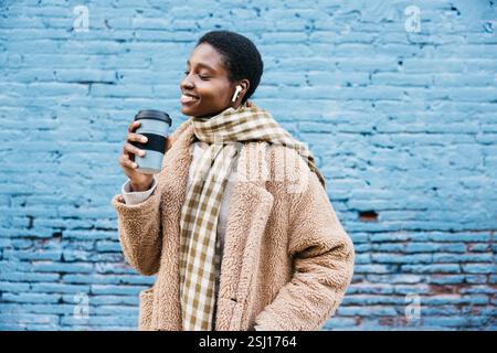 Stilvolle junge Frau, die einen warmen Mantel und Schal trägt, eine Einwegkaffeetasse hält und Musik mit kabellosen Ohrstöpseln hört Stockfoto