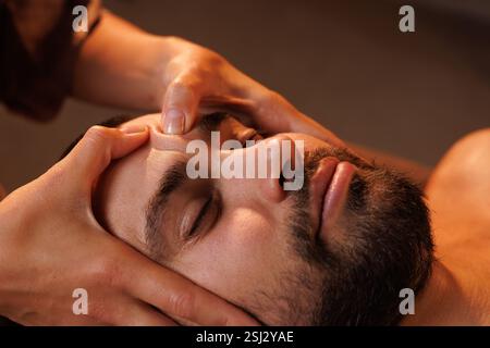 Nahaufnahme eines bärtigen Mannes, der eine entspannende Gesichtsmassage in einem Spa erhält. Warme Beleuchtung verstärkt die friedliche und therapeutische Atmosphäre dieses Selbstversorgers Stockfoto