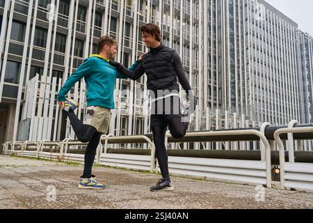 Zwei männliche Athleten dehnen die Beine zusammen, bevor sie in der Stadt laufen, lächeln und ihren gesunden Lebensstil genießen Stockfoto