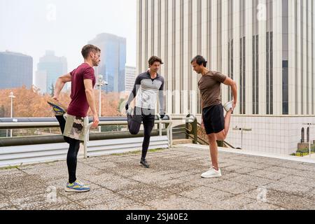 Drei Männer dehnen ihre Beine in einer lebhaften urbanen Umgebung und bereiten sich morgens auf ein belebendes Outdoor-Workout vor Stockfoto
