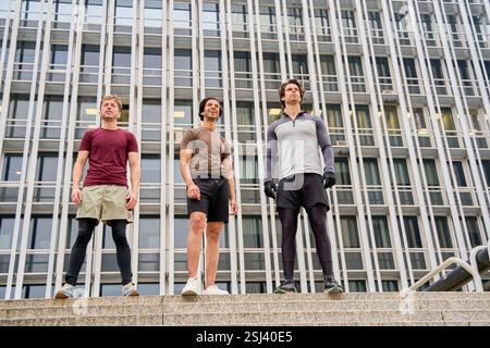 Drei männliche Athleten, die eine Pause vom Training im Freien machen, stehen auf Stufen mit einem großen modernen Gebäude dahinter Stockfoto