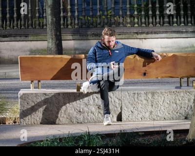 Ein Mann mit einer Rab-Jacke sitzt auf einer Bank aus Holz und Stein in der Nähe von One New Change, City of London, Großbritannien, vertieft in sein Handy Stockfoto