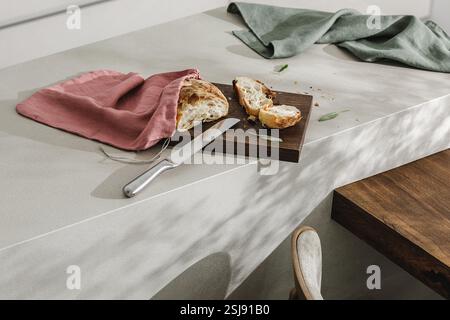 Geschnittenes Brot in Leinensack auf Holzbrett, frische Kräuter und rostfreies Destilliermesser. Küchenästhetisches Konzept Stockfoto