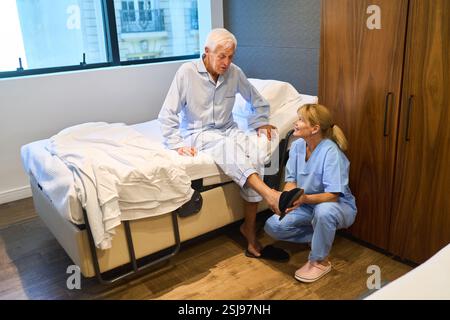 Älterer Mann, der auf dem Bett sitzt und ihm in einer Rehabilitationseinrichtung assistiert. Die unterstützende Szene veranschaulicht Pflege, Gesundheit und Erholung Stockfoto