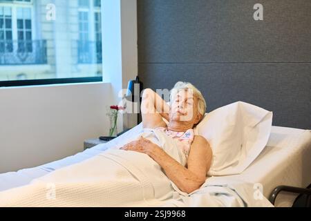Eine ältere Frau entspannt sich im Bett einer Rehabilitationseinrichtung, umgeben von modernen Möbeln. Stockfoto