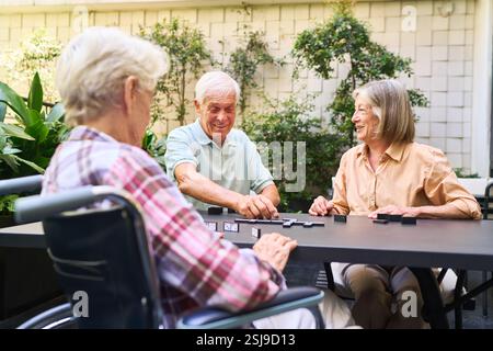 Ältere Menschen genießen ein Brettspiel im Freien, Lächeln und Begleitung in einer einladenden Gartenumgebung des Rehabilitationszentrums Stockfoto