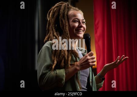 Frau mit Dreadlocks lächelt, während sie auf der Bühne eine Präsentation hält und ein Mikrofon hält. Begeistern Sie das Publikum mit ausdrucksstarken Gesten, die vor roten Vorhängen stehen Stockfoto