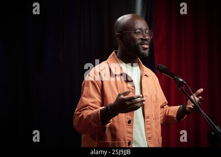 Afroamerikaner lächelt, während er während der Veranstaltung am Mikrofon spricht. Der Mann trägt eine orange Jacke und steht vor einem roten und schwarzen Hintergrund Stockfoto