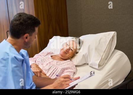 Ein medizinisches Fachpersonal kommuniziert mit einer älteren Frau, die in einem Bett einer Rehabilitationseinrichtung ruht, was medizinische Versorgung, Mitgefühl und Patientenversorgung symbolisiert Stockfoto